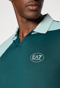 Zöld póló, textúrált anyagból, világoskék gallérral, az EA7 Emporio Armani logóval a bal mellrészen.