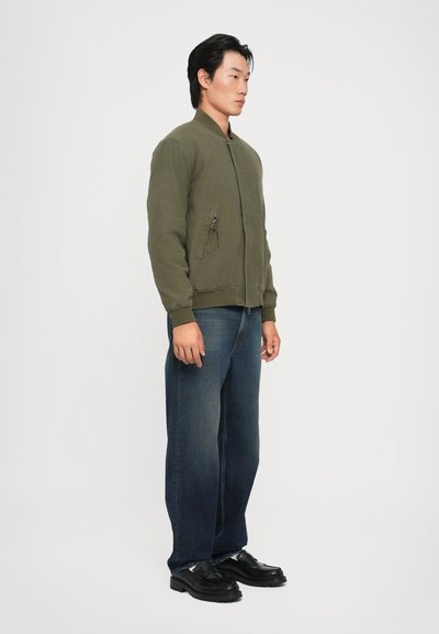 Olivgrön bomberjacka med framdragkedja, ribbade muddar och två sidofickor. Matchad med mörkblå raka jeans och svarta skor.