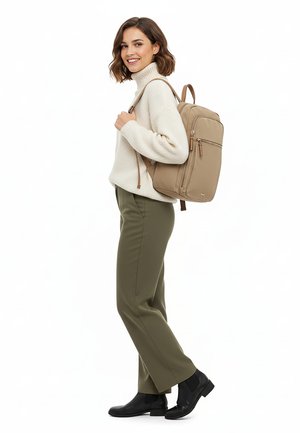 Kobieta w kremowym golfie i oliwkowych spodniach niosąca beżowy plecak, ubrana w czarne botki, uśmiechająca się i patrząca na bok.