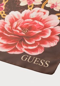 Hedvábný šátek s velkými růžovými květinami na hnědém pozadí, s řetězovým vzorem a logem "GUESS" v světle béžové. Hladký povrch.