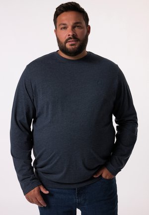 Uomo con barba che indossa una camicia blu scuro a maniche lunghe e jeans blu, in piedi con una mano in tasca davanti a uno sfondo uniforme.