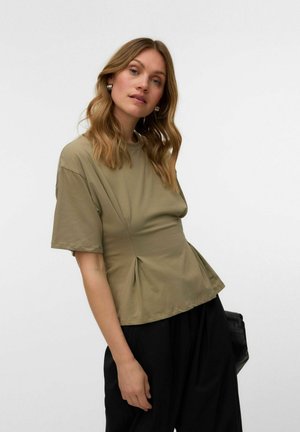 Femme aux cheveux blonds ondulés portant des boucles d’oreilles créoles dorées, un haut vert olive à manches courtes et un pantalon noir ample, tenant un sac noir.