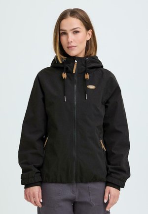 Kvinde iført en sort hættetrøje med lynlås, snore og forlommer, stående mod en ensartet lys baggrund.