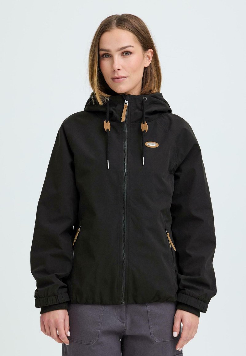 Femme portant une veste noire à capuche zippée avec cordons et poches avant, debout devant un fond clair uni.