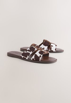 Pantoletten im Kuhmuster mit braunen Akzenten, ausgestattet mit zwei breiten Riemen, einer glatten Sohle und einer weichen Textur.