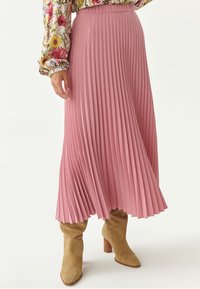 Plisseret midi-nederdel i blødt pink stof, med dybe vertikale plisseringer og et flagrende design. Parret med lysebrune, hælede støvler.