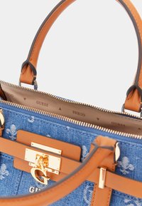 Denim handbag with brown leather accents, gold hardware, and a zip closure. Features interior with a repeating "Guess" logo pattern.