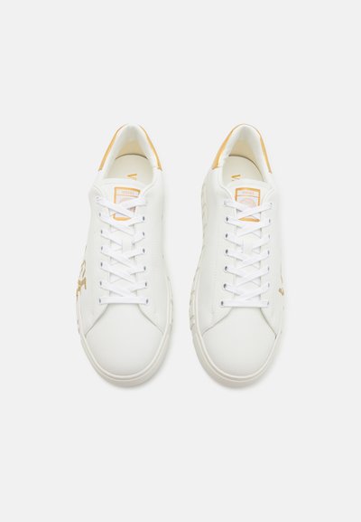 White synthetic sneakers with gold accents, round toe design, and flat laces. Branding visible on the side and tongue. Smooth texture.