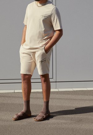 Mann trägt beige Tommy-T-Shirt und Shorts, kombiniert mit braunen Socken und Sandalen, steht auf einer gepflasterten Fläche an einer grauen Wand.