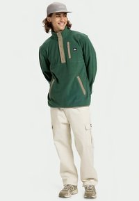 Roheline fliispullover kõrge kaelusega, nööbikaunistustega ja beeži tasku detailidega; koos koore värvi cargo-pükste ja beežide tossudega.