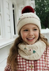 Strikket cremet hue med en rød pom-pom og dekorativ syning; cremet cowl med blomsterbroderi. Ternede rød og hvid frakke.