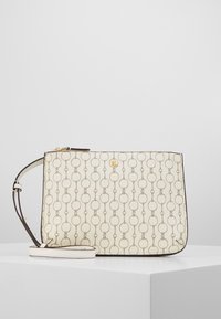 Krämfärgad axelremsväska med ett upprepande mönster av länkade ringar, guldlogotyp och justerbar rem, uppställd på en vit pelare mot en enkel bakgrund.