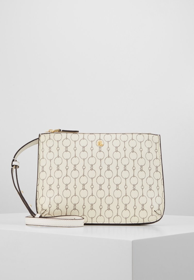 Krämfärgad axelremsväska med ett upprepande mönster av länkade ringar, guldlogotyp och justerbar rem, uppställd på en vit pelare mot en enkel bakgrund.