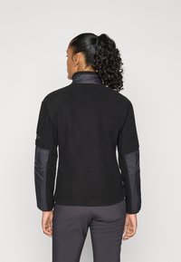 Vrouw met een krullende paardenstaart, gekleed in een zwarte fleece jack met gewatteerde elleboog- en kraag patches, staande tegen een effen lichte achtergrond.
