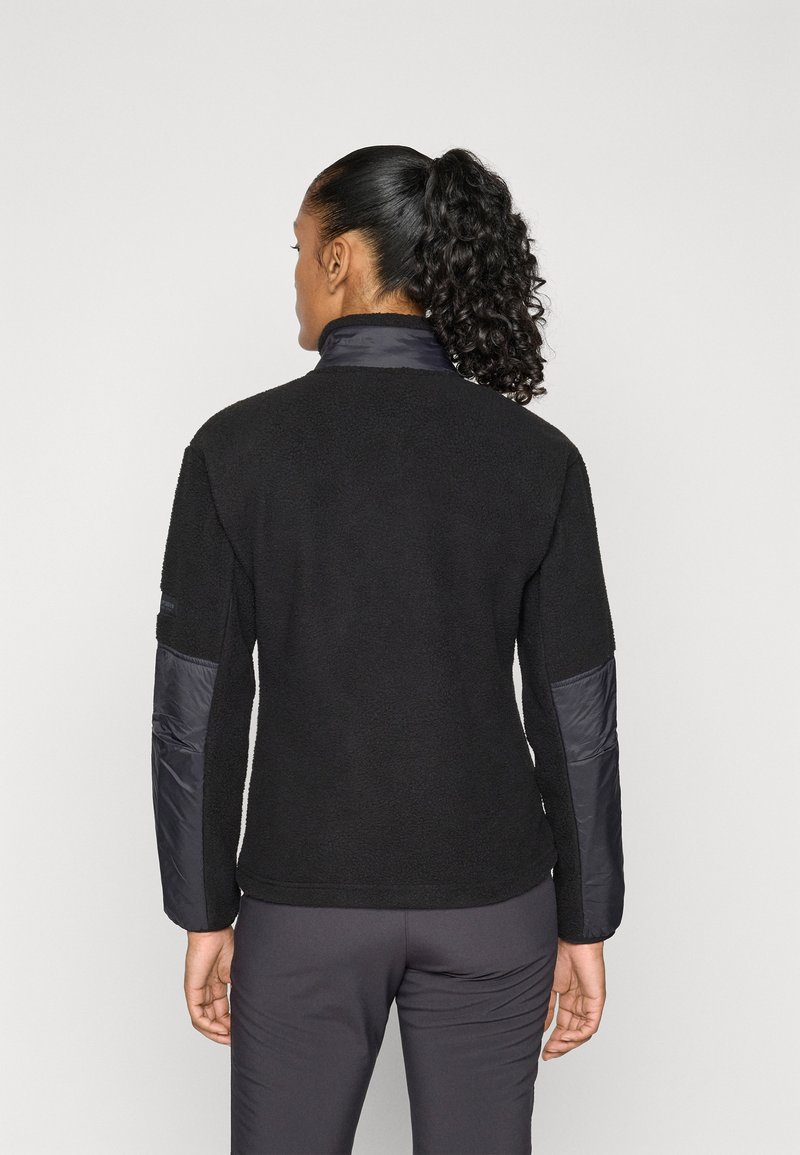 Frau mit lockigem Pferdeschwanz, die eine schwarze Fleecejacke mit gepolsterten Ellbogen- und Kragenpatches trägt, vor einem hellen, einfarbigen Hintergrund stehend.