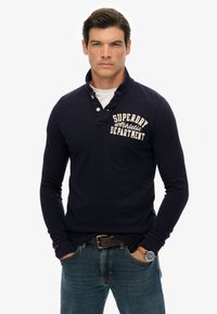 Polo de manga larga en color navy con cuello abotonado, que presenta un logotipo impreso en blanco en el pecho y una tela texturizada. Combinado con vaqueros azules.