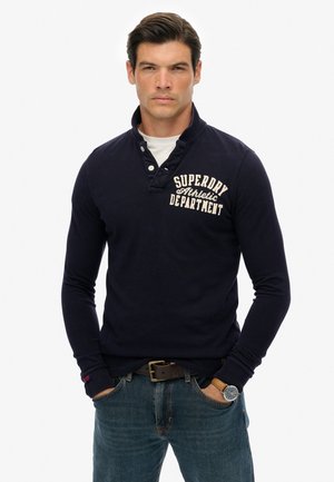 Marineblauwe polo met lange mouwen en een knoopsluiting bij de kraag, voorzien van een wit bedrukt logo op de borst en een gestructureerde stof. Gecombineerd met blauwe spijkerbroek.