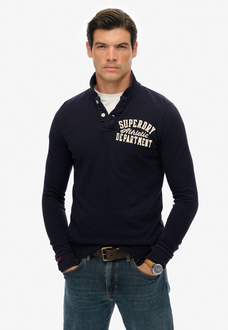 Polo de manga larga en color navy con cuello abotonado, que presenta un logotipo impreso en blanco en el pecho y una tela texturizada. Combinado con vaqueros azules.