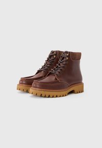 Botas de tobillo de cuero marrón con punta redonda, suelas de goma contrastantes y un cuello de gamuza marrón. Presenta ojales de metal y detalles de costura.