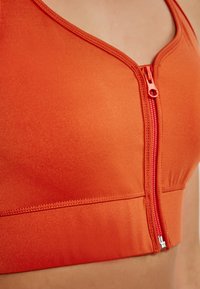 Sportovní podprsenka na zip v jasně oranžové barvě, vyrobená z hladkého materiálu. Obsahuje véčkový výstřih a kontrastní zip pro zapínání.