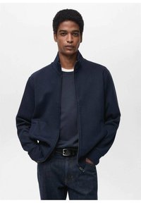 Giacca a zip navy in tessuto morbido con collo alto, dotata di tasche laterali e un fit rilassato, indossata sopra una camicia scura e jeans blu.
