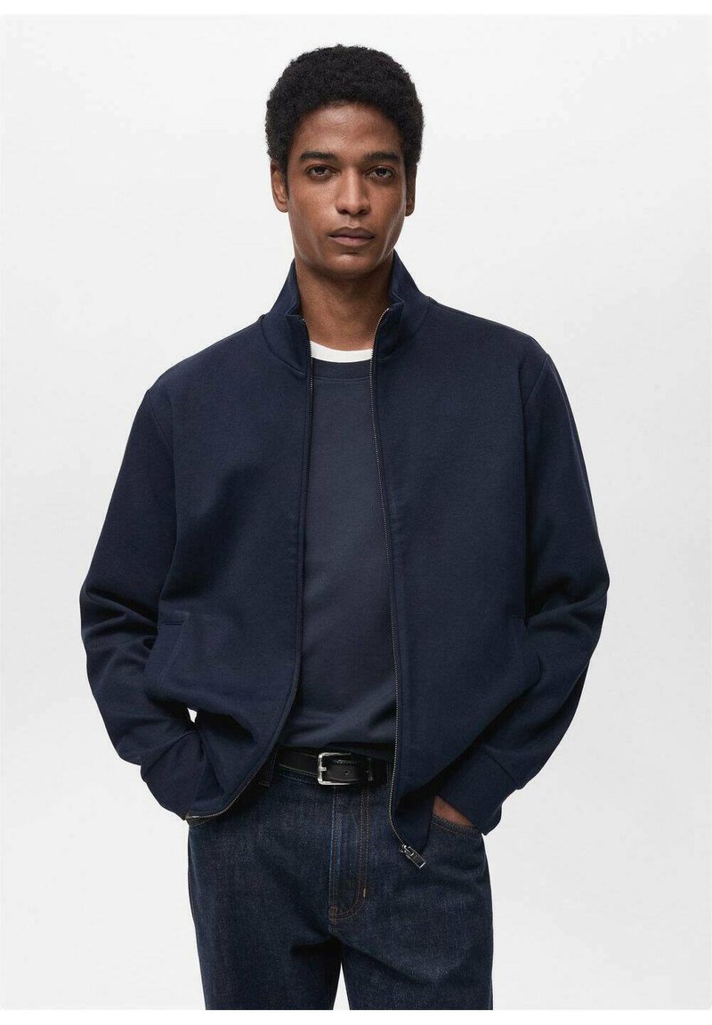 Giacca a zip navy in tessuto morbido con collo alto, dotata di tasche laterali e un fit rilassato, indossata sopra una camicia scura e jeans blu.