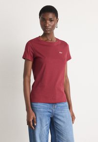Burgundy T-shirt with short sleeves, round neckline, and embroidered logo detail. Paired with light blue wide-leg jeans, showcasing a casual look.