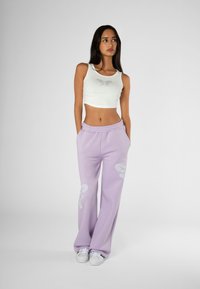 Bijela skraćena majica s detaljima od rhinestonea i lavanda trenirke s velikim bijelim grafikom. Model nosi bijele tenisice.