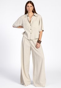 Chemise beige en lin à manches courtes, avec un devant �à boutons et deux poches, associée à un pantalon large assorti, présentant une texture douce.