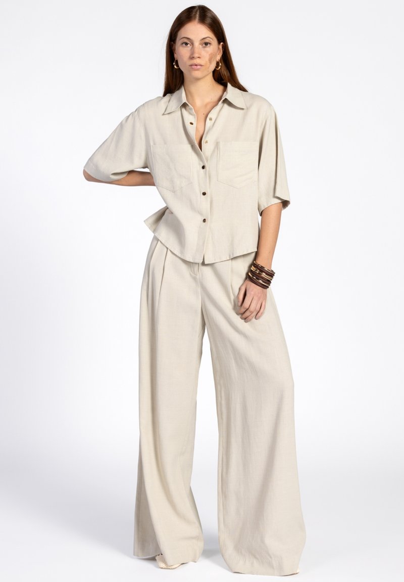 Chemise beige en lin à manches courtes, avec un devant à boutons et deux poches, associée à un pantalon large assorti, présentant une texture douce.