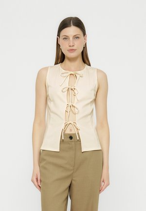 Femme portant un haut crème sans manches avec quatre nœuds devant et un pantalon beige taille haute, debout sur un fond clair uni.