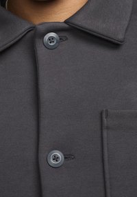 Ciemnoszara koszula z kołnierzykiem zapinanym na guziki, dwoma guzikami oraz małą kieszonką po lewej stronie, wykonana z gładkiego, teksturowanego materiału.