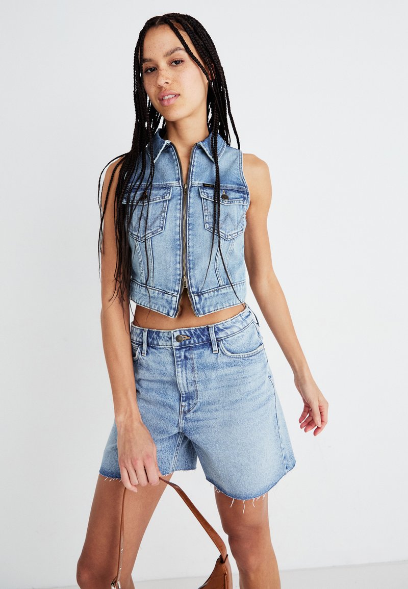 Denim-Crop-Weste mit Reißverschluss und Fronttaschen, kombiniert mit hochgeschnittenen Denim-Shorts. Hellblaue Farbe mit ausgefransten Saumdetails.