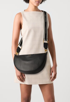 Zwarte lederen crossbody tas met een gebogen vorm, voorzien van een gouden logo en een gestreepte verstelbare schouderriem. Gemaakt om te dragen over een licht geruit jurkje.