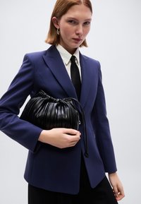 Μαύρη δερμάτινη τσάντα με πλισέ και κορδόνι, με απαλό φινίρισμα. Κρατιέται στο χέρι, συνδυασμένη με μπλε σακάκι, λευκό πουκάμισο και μαύρη γραβάτα.