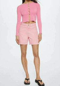 Top corto a maniche lunghe rosa a coste con dettagli a bottone e un'apertura a chiave; abbinato a pantaloni shorts in denim rosa chiaro. Sandali neri completano l'outfit.