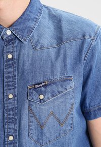 Camicia in denim a maniche corte di colore azzurro chiaro, con colletto a punta, chiusura con bottoni e taschino sul petto con un motivo a zigzag distintivo.