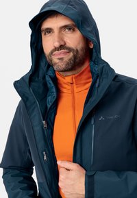 Mannen Vaude jas in donkerblauw met een capuchon, voorzien van een ritssluiting aan de voorkant, een oranje binnenlaag en een subtiele logo-detail op de borst.