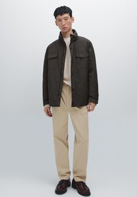 Giacca verde scuro con due tasche sul petto, indossata sopra un maglione crema, abbinata a pantaloni beige e scarpe stringate marroni. Design minimalista.
