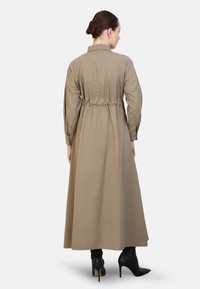 Lange, khaki-kleurige jurk met een elastische taille, lange mouwen en een overhemdkraag. Geaccentueerd met zwarte hoge hakken. Gladde textuur.