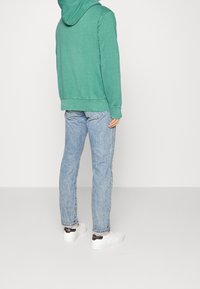 Mann mit einem grünen Kapuzenpullover, hellblauen Jeans und weißen Sneakers, der vor einem schlichten weißen Hintergrund steht, Rückansicht.