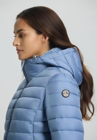 Femme aux longs cheveux bruns portant une veste matelassée bleu clair avec capuche et logo de marque sur le bras supérieur, tournée vers la gauche.