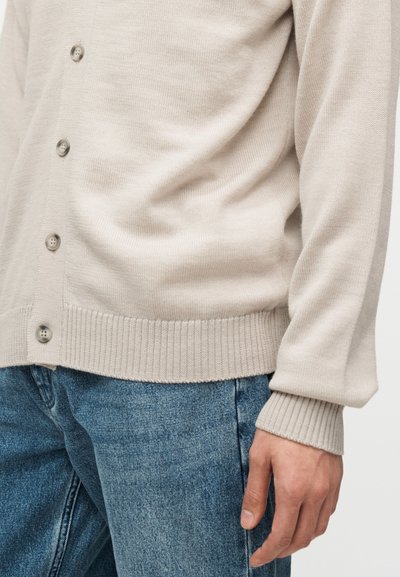 Μπεζ ζακέτα με κουμπιά, ριμπ μανσέτες και τελείωμα, φορεμένη πάνω από μπλε τζιν, δείχνοντας ένα χαλαρό εφαρμοστό στο κορμί και το χέρι του ατόμου.