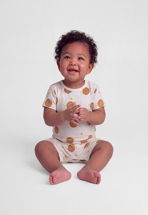 Bambino sorridente con capelli ricci seduto, indossa un abito bianco decorato con pianeti e stelle su uno sfondo semplice.