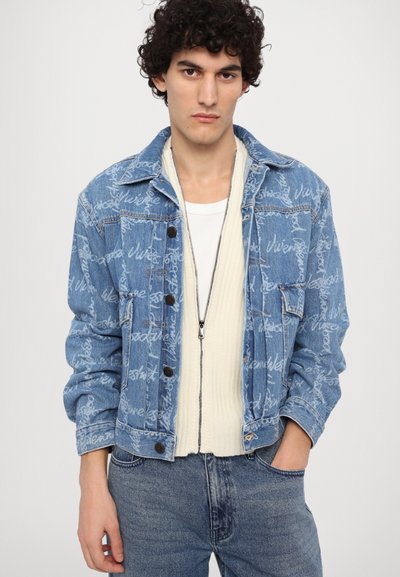 Denim jacket in light blue, featuring repeated white text pattern, button closure, chest pockets, and relaxed fit. Worn over a cream sweater.