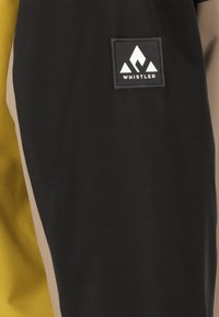 Svart skjorta med en gul sidopanel och en beige accent; inkluderar en fyrkantig tygetikett med en vit logotyp som säger "WHISTLER."