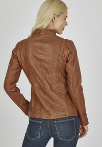 Veste en cuir marron au design structuré, avec des coutures verticales et un col cheminée. Portée avec un jean bleu marine.