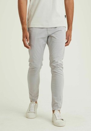 Mand iført lysegrå slim-fit bukser, hvide sneakers og en hvid skjorte mod en ensfarvet lys baggrund.