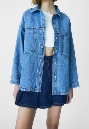 Jeune personne portant une veste en jean bleu clair oversize, un crop top blanc et une jupe plissée en jean bleu foncé sur fond uni.