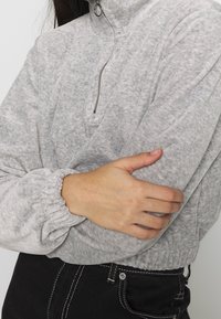 Grå, texturerad fleecepullover med en halvhög krage, elastiska muddar och en åtsittande midja, kombinerad med svarta jeans med kontrasterande sömmar.